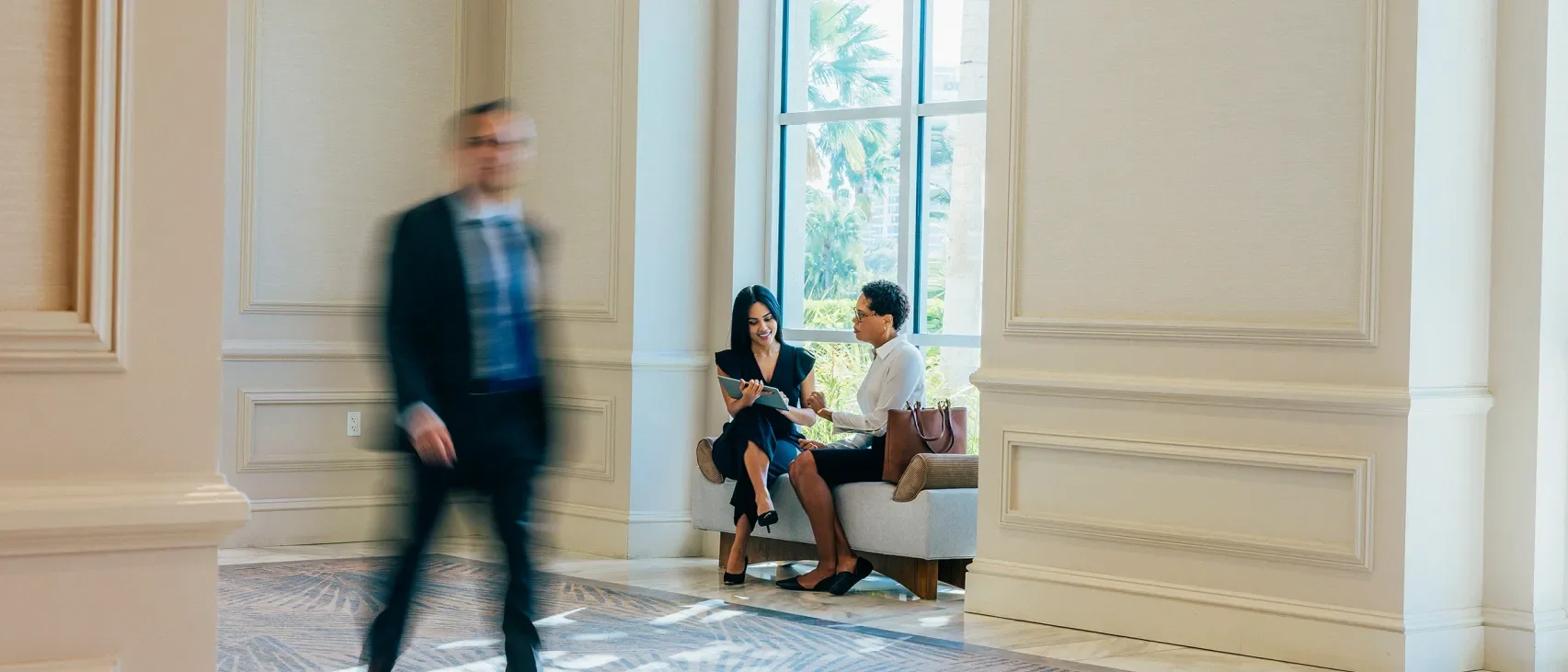 Man in a suit walks past two women in business attire looking at a document